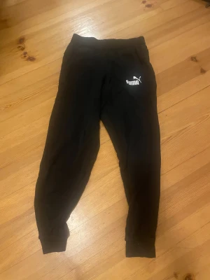 Svarta mjukisbyxor från Puma M - Svarta mjukisbyxor från Puma i storlek M. Byxorna har resår i midjan och vid bensluten samt en vit Puma-logga på låret. Perfekta för chill eller träning, tillverkade i mjukt material för maximal komfort.