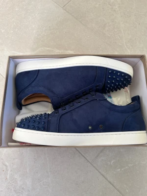 Mörkblå Christian Louboutin sneakers - Säljer ett par mörkblå sneakers från Christian Louboutin med snygga nitar på tån och sidorna. Skorna har en platt vit sula med röd undersida, snörning och är tillverkade i mocka med läderfoder. Perfekt för dig som gillar att sticka ut med coola detaljer.