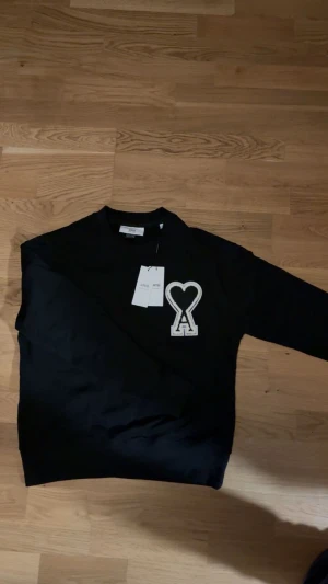 Svart sweatshirt från Ami med hjärtdetalj - Hel ny svart sweatshirt från Ami Paris med en unik logga. Materialet är mjuk bomull och logotypen Ami syns även baktill i nacken. Perfekt för en clean och stilren look. Hör av dig vid minsta funderingar!!!