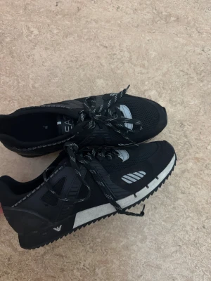 Svarta sneakers från Emporio Armani - Snygga svarta sneakers från Emporio Armani med vita och grå detaljer. Skorna har en sportig design med mesh och syntetmaterial, samt mönstrade snören med EA7-logga. Sulan är vit med svarta inslag och har en räfflad undersida för extra grepp.