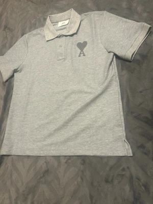 Grå polo från Ami med hjärtdetalj - Snygg grå pikétröja från Ami Alexandre Mattiussi med klassisk krage och knappar framtill. På bröstet finns Ami:s ikoniska hjärta med bokstaven A broderat i mörkgrått. Mjuk bomullskvalitet och korta ärmar gör den chill och enkel att bära.