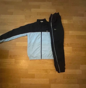 Lacoste blå och svart träningsset - Snyggt träningsset från Lacoste med jacka och byxor i svart och ljusblått. Jackan har dragkedja framtill, elastisk midja och vit Lacoste-logga på ryggen. Byxorna har vit piping längs benen och klassisk Lacoste-krokodil på höften. Perfekt för sportiga dagar.