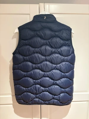 Dunväst Peak Performance - Snygg mörkblå dunväst från Peak Performance med vågformat quiltat mönster. Västen har hög krage, dragkedja framtill och två sidofickor med dragkedja. Perfekt för lager-på-lager och håller dig varm under kyliga dagar. Ny pris 1599 mitt pris 729! ( passar S-M) 