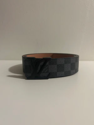 Rutig svart bälte - Snyggt bälte med klassiskt rutigt mönster i svart och grått. Bältet har en bred form och en stor logga i svart metall som spänne. Tillverkat i exklusivt skinn med lyxig känsla och detaljerad finish. Perfekt för dig som vill ha en statement-accessoar.