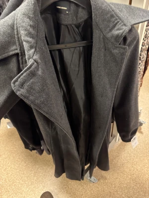 Mörkgrå kappa från Vero Moda - Snygg mörkgrå kappa från Vero Moda med breda slag och klassisk krage. Jackan har en rak och stilren siluett, perfekt för dig som gillar enkel och clean look. Materialet är mjukt och känns lyxigt, med helsvart foder på insidan. 