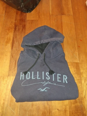 Mörkblå Hollister hoodie - Säljer en mörkblå hoodie från Hollister med stor logga och text på bröstet. Tröjan har huva med snörning och är tillverkad i mjuk bomullsmix. Perfekt för chill dagar och har en klassisk streetstil. Hör gärna av er vid funderingar eller prisförslag!!