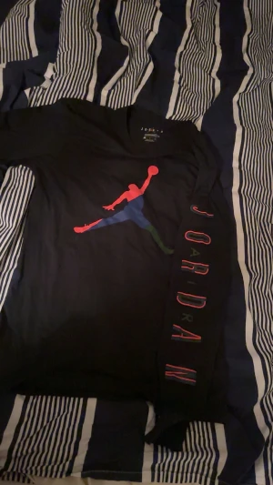 Svart långärmad Jordan tröja - Svart långärmad tröja från Jordan med ikonisk Jumpman-tryck i rött, blått och grönt på bröstet. På ena ärmen står JORDAN i färgglada bokstäver. Tröjan har rund hals och är gjord i mjuk bomull, perfekt för dig som gillar sportig streetstyle. Säljer för aldrig använt den‼️