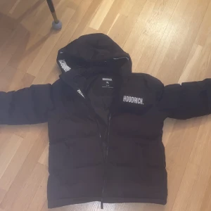 Svart pufferjacka från Hoodrich XL - Svart pufferjacka från Hoodrich i storlek XL med huva och vita logotexter på bröstet, huvan och ärmarna. Jackan har dragkedja framtill och är quiltad för extra värme. Perfekt för kalla dagar och har en modern, loose passform. Den har ett litet hål k fickan men inget man märker eller kan se 