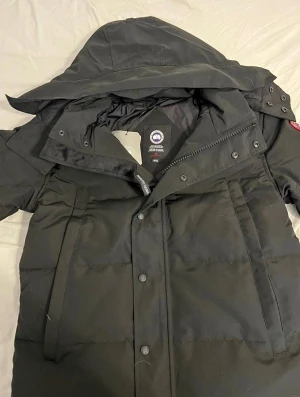 Svart dunjacka från Canada Goose - Svart dunjacka från Canada Goose med huva och klassisk logopatch på ärmen. Jackan har dragkedja och knappar framtill, samt flera praktiska fickor. Puffad design och justerbar huva gör den perfekt för kalla dagar. Snygg och funktionell för dig som vill ha värme och stil.