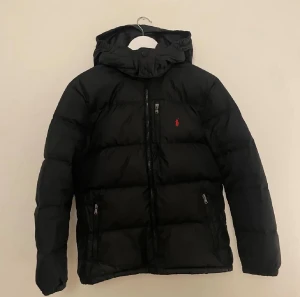 Svart pufferjacka från Polo Ralph Lauren - Svart pufferjacka från Polo Ralph Lauren med huva och dragkedja framtill. Jackan har två sidofickor med dragkedja och en liten röd Polo-logga på bröstet. Perfekt för kalla dagar och har en clean, sportig look. Det står Xl men om man är runt 165-180 passar den 