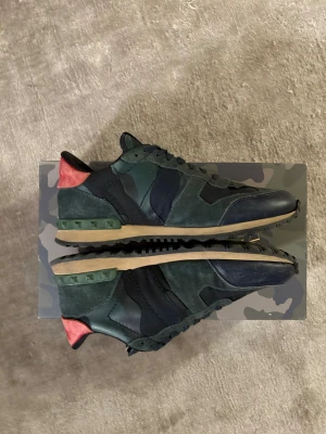 Valentino Rockrunner sneakers camo - Säljer ett par Valentino Rockrunner sneakers i mörkgrönt, svart och marinblått med röda detaljer vid hälen. Skorna har camouflagemönster, snörning och platt sula. Materialmix av mocka, skinn och textil. Ikoniska nitar på hälen och klassisk Valentino-logga på plösen.
