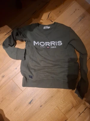 Olivgrön sweatshirt från Morris - Snygg olivgrön sweatshirt från Morris i storlek S. Tröjan har rund hals, långa ärmar och vit logotyp med brittisk flagga på bröstet. Tillverkad i mjuk bomull med ribbade muddar vid ärmslut och nederkant. Perfekt för chill dagar och streetwear.