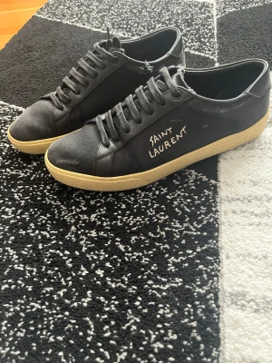 Svarta Saint Laurent sneakers strl 41 - Svarta sneakers från Saint Laurent med handskriven logga på sidan. Snygg kontrast med beige sula och svarta snören. Tillverkade i Italien med en mix av skinn och textil, rund tå och platt sula. Perfekt för dig som gillar stilrena designer med edge. Ända defekterna är att sulan är lite gulare än den ska och en av slitningen (modellen ska ha slitningar) har blivit större än alla andra. Men fortfarande skitsnygga! Passa på att bli grisch inför vintern. Dustbag medföljer
