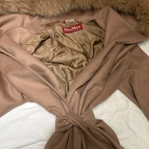 Beige Max Mara kappa med päls - Snygg beige kappa från Max Mara Studio med lyxig pälsdetalj vid kragen och knytband i midjan. Insidan är fodrad i champagnefärgad satin och kappan har en elegant omlottstil. Perfekt för dig som vill ha en classy och tidlös look. köptes mellan 10 000 - 11 000:- . Äkta kashmir säljs för 3000:- 