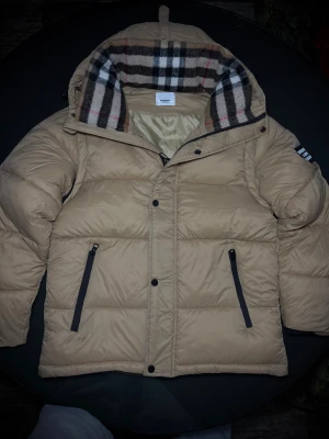 Beige pufferjacka från Burberry - Snygg beige pufferväst från Burberry med ikoniskt rutigt foder i huvan. Västen har svarta dragkedjor på fickorna, tryckknappar och stor Burberry-logga på insidan. Perfekt för dig som vill ha en clean och klassisk look med premiumkänsla.