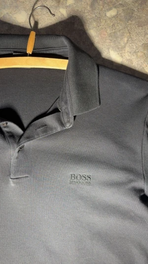 Svart piké från Hugo Boss, storlek M - Svart pikétröja från Hugo Boss med klassisk krage och knappar framtill. Diskret broderad logga på bröstet. Tillverkad i mjuk bomull som känns skön mot huden. Perfekt för en clean och stilren look.