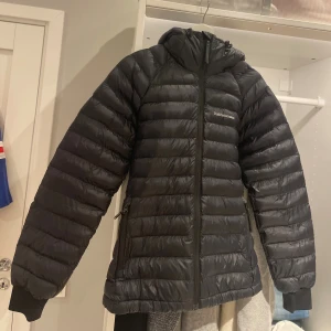 Svart dunjacka Peak Performance - Svart dunjacka från Peak Performance med huva och två sidofickor med dragkedja. Jackan har quiltad design, ribbade muddar och Peak Performance-logga på bröstet samt ett brett band med logga på huvan. Perfekt för kalla dagar och snygg till streetwear. In princip helt nytt skick. 