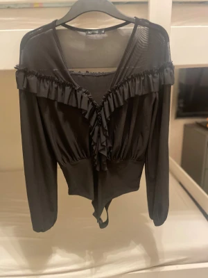 Svart volang body från NLY One  - Svart body från NLY One i storlek XS med transparent mesh upptill och långa ärmad. Toppen har snygga volangdetaljer längs v-ringningen och axlarna, vilket ger en cool och feminin vibe. Perfekt att styla med höga jeans eller kjol för en trendig look.