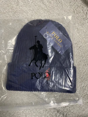 Marinblå ribbstickad mössa Polo Ralph Lauren - Snygg marinblå ribbstickad mössa från Polo Ralph Lauren med klassisk röd broderad polospelare framtill. Mössan har uppvikt kant och är tillverkad i mjukt material som håller dig varm och stilren under kalla dagar. Helt äkta direkt från Polo.  Skickas samma dag eller nästa dag 🤩☺️
