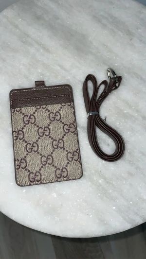  brun plånbok med axelrem - Snygg plånbok i brunt skinn och beige canvas med klassiskt monogram mönster. Kommer med matchande brun axelrem i skinn och har fack för kort samt ett fönster för ID på baksidan. Perfekt för att bära det viktigaste med stil.