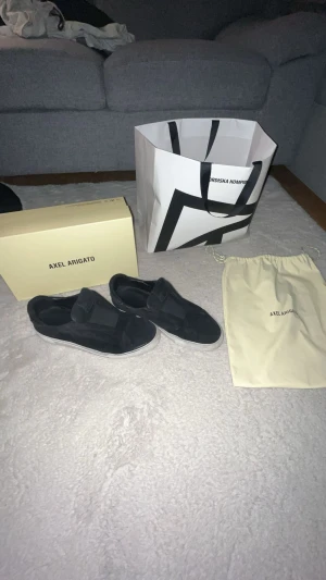 Svarta sneakers från Axel Arigato - Svarta sneakers från Axel Arigato med stilren design och platt vit sula. Skorna är tillverkade i mocka och har elastiska paneler istället för snörning, vilket ger en clean och modern look. Perfekta för dig som gillar minimalistisk stil och premiumkänsla.