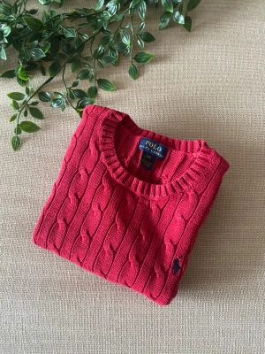 Röd kabelstickad tröja Polo Ralph Lauren - Snygg röd kabelstickad tröja från Polo Ralph Lauren med rund hals och klassisk marinblå logga broderad på bröstet. Tröjan har ribbade muddar vid ärmslut och nederkant, och är tillverkad i mjuk bomull. Perfekt för dig som gillar preppy stil.