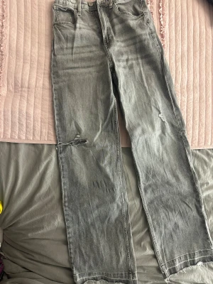 Grå raka jeans med slitningar - Säljer ett par grå jeans med raka ben och slitna detaljer på knäna. Jeansen har klassisk femficksdesign, normal midja och råa kanter vid bensluten. Perfekta för en avslappnad och trendig look.