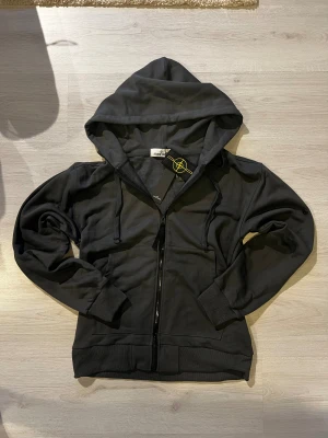 Mörkgrå hoodie från Stone Island - Svart hoodie från Stone Island med klassisk logopatch på ärmen. Tröjan har dragkedja framtill, huva med snörning och två fickor. Tillverkad i mjuk bomull och har ribbade muddar vid ärmslut och nederkant. Perfekt för dig som gillar streetwear och snygga detaljer.