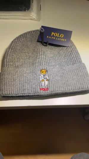 Grå ribbad mössa Polo Ralph Lauren - Snygg grå ribbstickad mössa från Polo Ralph Lauren med broderad nalle framtill och röd POLO-text. Mjuk och varm, perfekt för kyliga dagar. Materialet är 100% akryl och mössan har en klassisk uppvikt kant.