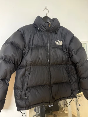 Svart pufferjacka från The North Face - Säljer en svart pufferjacka från The North Face i storlek M. Jackan har hög krage, dragkedja framtill och snörning vid nederkant. Klassisk logga på bröstet och ryggen. Perfekt för kalla dagar med sin tjocka, quiltade design och stilrena look.