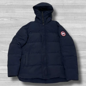 Canada Goose jacka - Säljer en fet marinblå dunjacka från Canada Goose i storlek XXL men passar även XL och L. Jackan har en klassisk pufferstil med huva, svart innerfoder och dragkedja framtill. Perfekt för kalla dagar och har den ikoniska Canada Goose-loggan på ärmen.