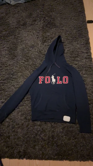 Mörkblå hoodie från Polo Ralph Lauren - Snygg mörkblå hoodie från Polo Ralph Lauren med stor POLO-logga i rött och vitt på bröstet. Tröjan har huva med snörning, känguruficka och långa ärmar. Perfekt för chill dagar och streetwear-stil.