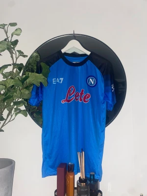 Napoli blå fotbollströja EA7 - Säljer en officiell Napoli fotbollströja från EA7 med korta ärmar. Tröjan är blå med svarta axlar och har tryck med klubbmärke, sponsor och SSCN på ryggen. Materialet är lätt och andas, perfekt för match eller träning.