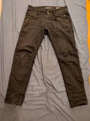 Svarta Replay Anbass slim jeans 27/30 - Svarta jeans från Replay, modell Anbass Slim Fit. Klassisk femficksdesign med dragkedjegylf och snygga detaljer vid fickorna. Jeansen har en smal passform och är tillverkade i ett stretchigt bomullsmaterial för extra komfort. Nedkortade därför L30 ursprung L32