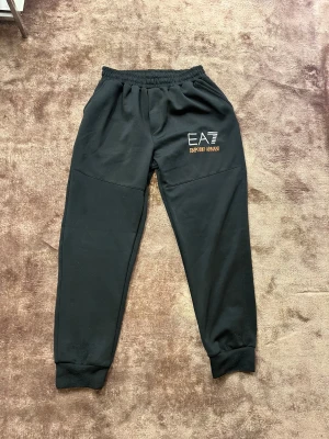 Svarta mjukisbyxor EA7 Emporio Armani - Svarta mjukisbyxor från EA7 Emporio Armani med elastisk midja och ribbade muddar vid bensluten. Byxorna har en stilren design med logotyptryck i vitt och guld på ena benet. Perfekta för chill eller träning, mjukt material och relaxed passform.