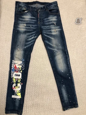 DSQUARED2 jeans med färgstänk och tryck - Snygga DSQUARED2 jeans i mörkblå tvätt med slitningar och vita färgstänk. Ena benet har stort färgglatt tryck med texten 'ICON' och DSQUARED2. Klassisk femficksmodell, slim fit och tillverkade i bomull. Perfekt för dig som gillar streetstyle och statement-plagg.