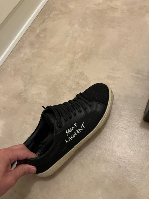 Svarta Saint Laurent sneakers - Svarta sneakers från Saint Laurent med vit logga på sidan och vit sula. Skorna har snörning och en stilren, rund tå. Materialet är skinn och textil, och medföljer gör originalkartong och dustbag. 