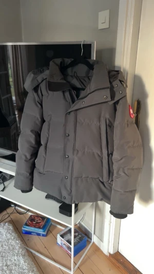 Mörkgrå pufferjacka XS - Säljer en mörkgrå pufferjacka i storlek XS. Jackan har huva, knappar och dragkedja framtill samt den klassiska röda loggan på ärmen. Perfekt för kalla dagar med sin tjocka och quiltade design.