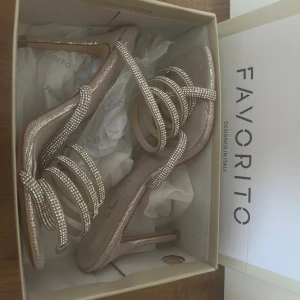Glittriga sandaler med spiralremmar - Säljer ett par sandaler från Favorito med höga klackar och spiralformade remmar dekorerade med glittrande stenar. Skorna är i en ljus beige ton med silverdetaljer och har öppen tå samt fyrkantig sula. Perfekta för att sticka ut med en riktigt glam look.