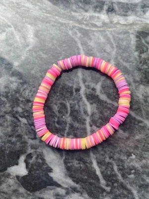 Färgglatt pärlarmband i pastell💗 - Säljer ett handgjort armband med platta pärlor i rosa, lila och ljusgult. Armbandet har ett randigt mönster och är elastiskt, vilket gör det enkelt att ta på och av. Perfekt för dig som gillar färg och vill ha något unikt på handleden.