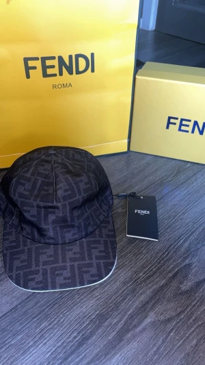 Fendi keps - Exklusiv svart keps från Fendi med ikoniskt FF-monogram över hela ytan. Kepsen har böjd skärm med vit kant och justerbar passform. Tillverkad i ett slitstarkt polyestertyg och levereras med originalpåse, box och taggar.