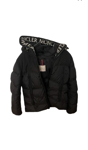 Svart Moncler dunjacka - Svart dunjacka från Moncler med stor huva och vit logotyptext längs kanten.  Nypris 11200 