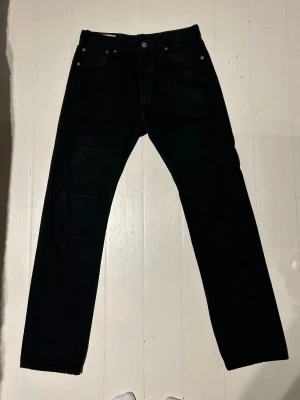 Svarta Levi's 501 W30 L32 - Svarta Levi's 501 Premium jeans med klassisk rak passform och femficksdesign. Jeansen är tillverkade i slitstarkt bomullstyg och har snygga kopparfärgade nitar samt den ikoniska läderpatchen bak i midjan. Perfekta för en clean och tidlös look.