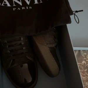 Svarta sneakers från Lanvin - Snygga svarta sneakers från Lanvin med glansig tå och snörning. Perfekta för en stilren look. Skorna kommer i originalförpackning med en tygpåse för förvaring. Tänk på att skorna är stora i storlek även om det är storlek Uk6 passar dom dessutom storlek 40-43 utan problem!
