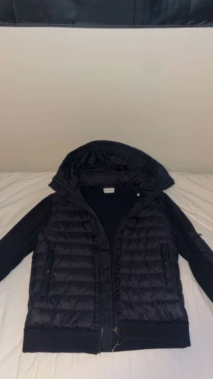 Mörkblå pufferjacka från Moncler - Säljer en mörkblå pufferjacka från Moncler med stickade ärmar och huva. Jackan har två sidofickor med dragkedja, ribbade muddar och klassisk Moncler-logga på ärmen. Materialmix av polyester och stickad textil ger en schysst look. Perfekt för dig som gillar streetstyle.