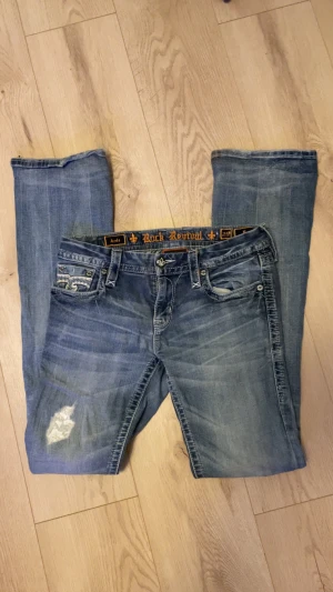  Rock Revival bootcut jeans - Snygga blå jeans från Rock Revival med bootcut passform. Jeansen har slitna detaljer och broderade bakfickor med dekorativa knappar. Klassisk femficksmodell med låg midja. Säljer pga dem var lite stora
