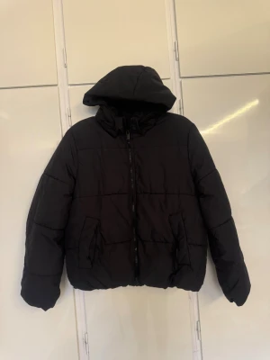Svart pufferjacka med huva H&M - Säljer en svart pufferjacka med huva och dragkedja framtill. Jackan har två stora fickor på sidorna och är quiltad för extra värme. Perfekt för kalla dagar och har en loose passform som ger en chill vibe.