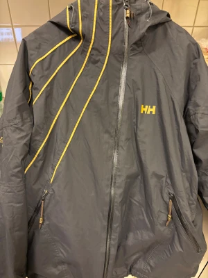 Svart och gul vindjacka Helly Hansen XL - Svart vindjacka från Helly Hansen i storlek XL med gul insida och detaljer. Jackan har huva, dragkedja framtill och två fickor med dragkedja. Snygga gula linjer på framsidan och HH-logga på bröstet och insidan. Perfekt för dig som gillar sportig stil.