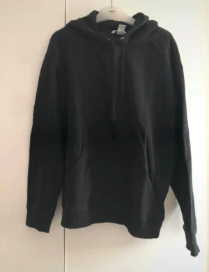 Svart hoodie med huva och ficka - En klassisk svart  hoodie med  huva och snörning, samt stor magficka framtill. Tröjan har lång ärm och en avslappnad passform. Perfekt för att slänga på sig när det är lite kyligt ute eller som ett stilrent basplagg i garderoben