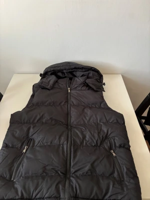 Svart dunväst med huva  - Svart dunväst med avtagbar huva och två fickor med dragkedja framtill. Västen har quiltad design och hel dragkedja i mitten. Perfekt för lager-på-lager och håller dig varm under kyliga dagar. Snygg och enkel att matcha med olika outfits. Helt ny.